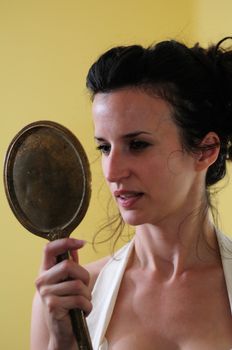 A young woman gazes at her reflection in an antique hand mirror against the background of a yellow wall.