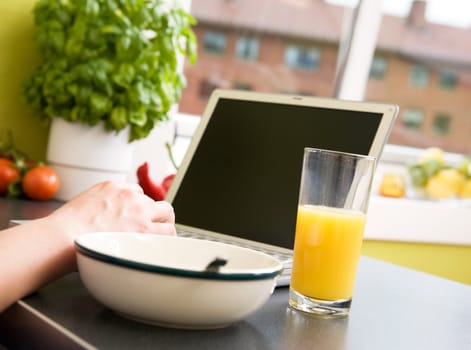 A computer in the kitchen with a bowl of soupr or cereal and orange juice - shallow depth of field with focus on juice and hand