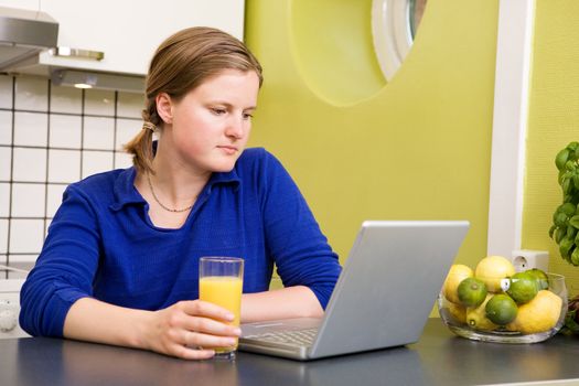 A young woman reads the morning news on a laptop while drinking orange juice