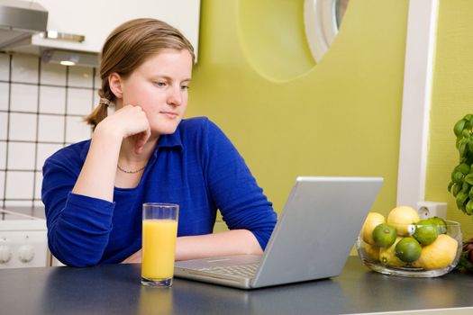 A young woman reads the morning news on a laptop while drinking orange juice