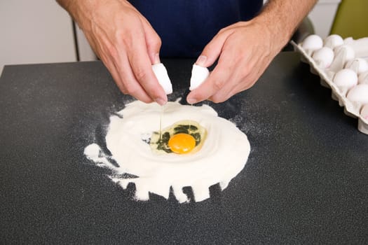 A young man making pasta at home in an apartment kitchen - Cracking an egg into the flour to be mixed by hand on the counter;