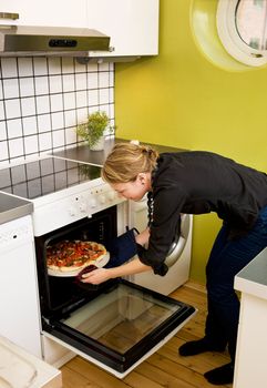 A young woman takes a fresh pizza out of the oven in her apartment kitchen.