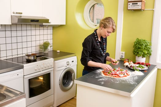 A young female in an apartment kitchen making an itialian style pizza.