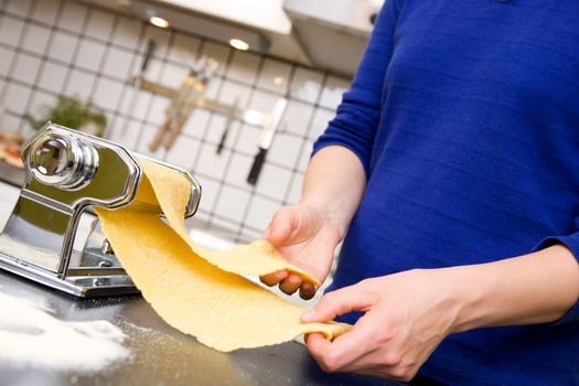 A young female stretching out pasta over the counter from a manual pasta machine at home in the kitchen.