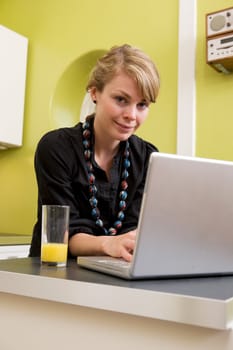 A young woman uses the computer in the kitchen while enjoying a glass of juice. The model is looking at the viewer and smiling.