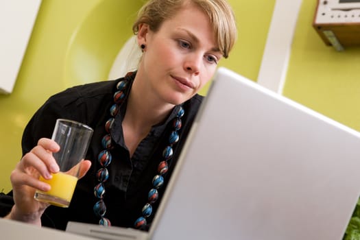 A young woman uses the computer in the kitchen while enjoying a glass of juice. The model is looking at the computer.