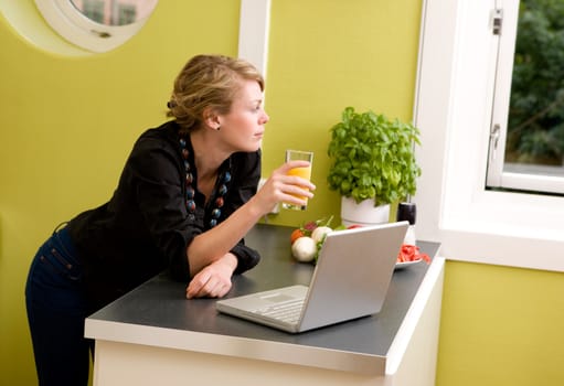 A woman using a laptop in the kitchen - looking out the window as though she is thinking about someone or something.