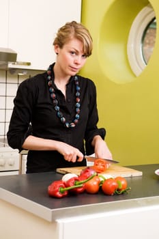 A female in her apartment kitchen slicing tomatoes and looking at the camera with a slight smile.