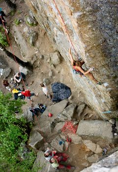 An eager female climber on a steep rock face looks for the next hold - viewed from above.  Shallow depth of field is used to isolated the climber.  A belayer and other climbers look on in anticipation.