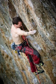 A male climber, viewed from above, climbs a very high and steep crag.