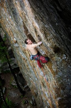 A male climber, viewed from above, climbs a very high and steep crag. 