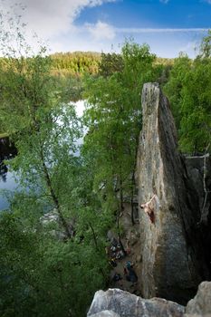 A male climber, viewed from above, climbs a very high and steep crag. A beautiful lake and landscape create a breathtaking backdrop.