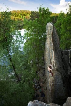 A male climber, viewed from above, climbs a very high and steep crag. A beautiful lake and landscape create a breathtaking backdrop.