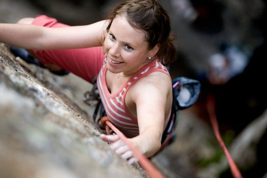 A female climber on a steep rock face viewed from above with the belayer in the background.  The climber is smiling at the camera. Shallow depth of field is used to isolated the climber.