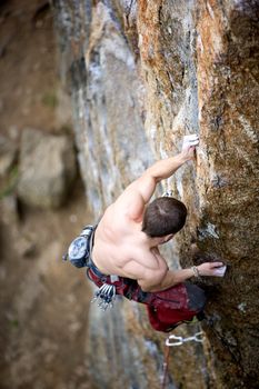 A male climber, viewed from above, climbs a very high and steep crag. Shallow depth of field is used to isolate the climber with focus on the hands and head