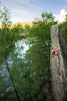 An eager female climber on a steep rock face looks for the next hold. Breathtaking scenery including a lake and forest are in the background. Shallow depth of field is used to isolated the climber with the focus on the head.