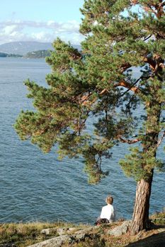 An eldery woman sitting on the rocks by the ocean under a tree.