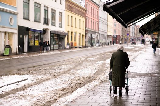 An old woman walking outdoors in the winter with a few snow crystals visible in the air. - Focus is on the woman with the background thrown out of focus.