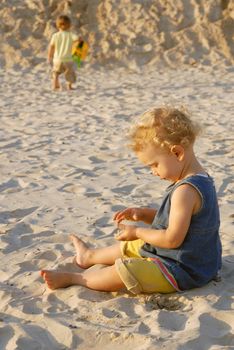 Little girl playing on the beach with sand. A boy in his back out of focus in the background.