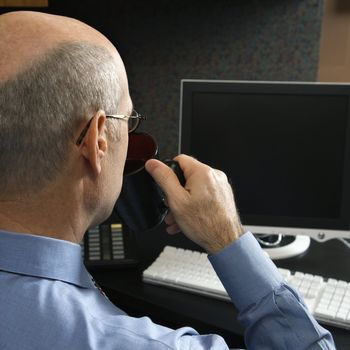 Back view of Caucasian middle-aged businessman drinking from coffee cup sitting at desk looking at computer.