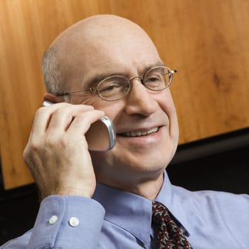 Smiling Caucasian middle-aged businessman sitting in office talking on cellphone.