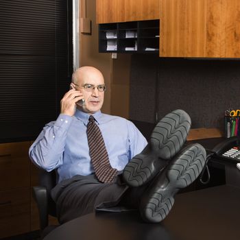 Caucasian middle-aged businessman in office sitting with feet on desk talking on cellphone.