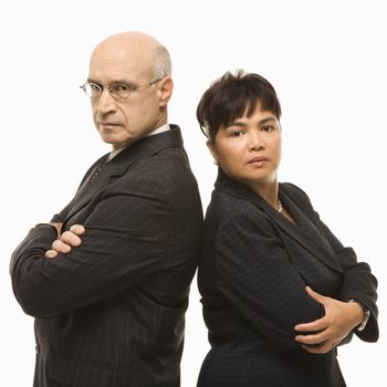 Caucasian middle-aged businessman and Filipino businesswoman standing back to back with arms crossed looking serious.