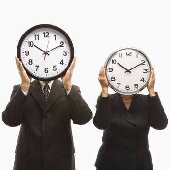 Caucasian middle-aged businessman and Filipino businesswoman holding clocks in front of their heads standing against white background.