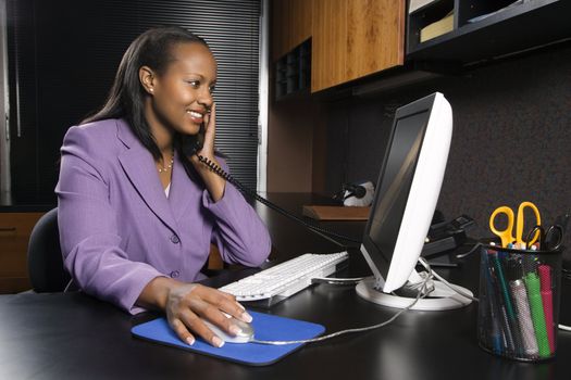 African-American young adult smiling business woman talking on phone and working at computer in office.