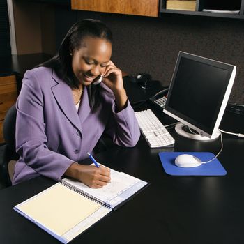 High angle view of African-American young adult business woman talking on cell phone and writing in planner in office.