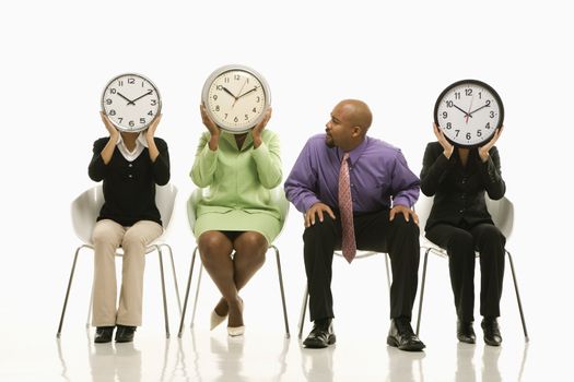 Businesswomen sitting holding clocks over faces while African-American businessman looks on.