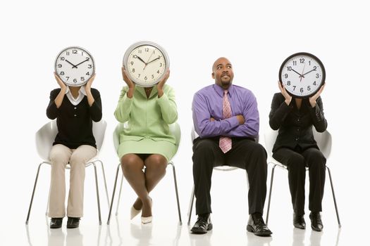 Businesswomen sitting holding clocks over faces while African-American businessman looks on.