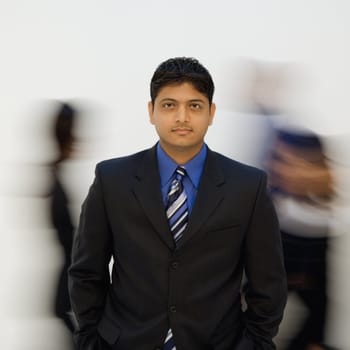 Indian businessman standing with hands in pockets while others walk by.