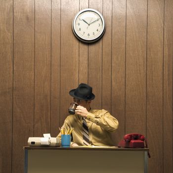 Caucasion mid-adult retro businessman wearing fedora sitting at desk drinking coffee.