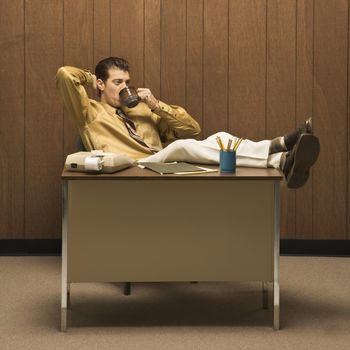 Caucasion mid-adult retro businessman sitting with feet propped on desk leaning back with hand behind head drinking coffee.