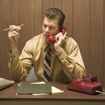 Caucasion mid-adult retro businessman sitting at desk talking on telephone.