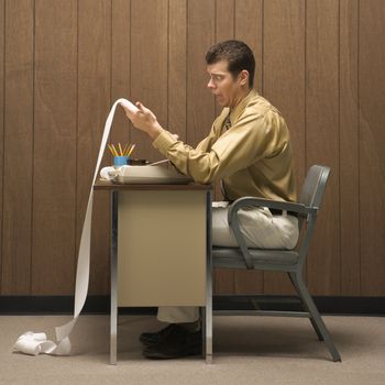 Caucasion mid-adult retro businessman sitting at desk looking with shocked expression at long adding machine printout that reaches all the way to the floor.