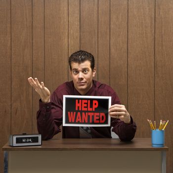 Caucasion mid-adult retro businessman sitting at desk holding help wanted sign with pleading expression.