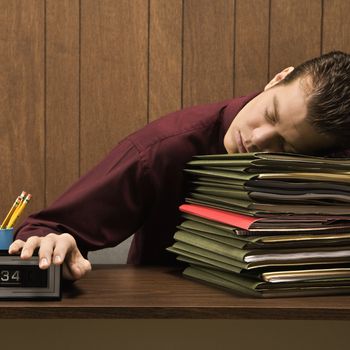 Caucasion mid-adult retro businessman sitting at desk with head down sleeping on a tall stack of folders.