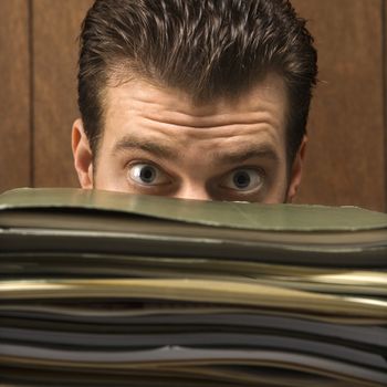 Caucasion mid-adult retro businessman peering with wide eyes over a tall stack of folders.