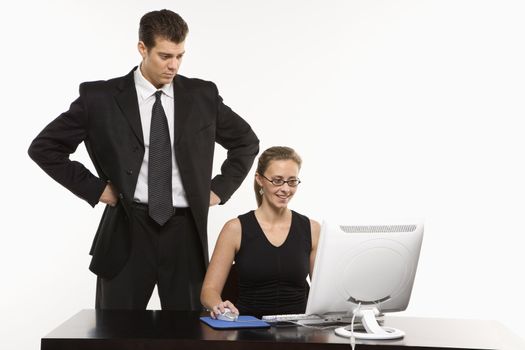 Caucasian mid-adult man with hands on hips looking over shoulder of woman sitting at computer.