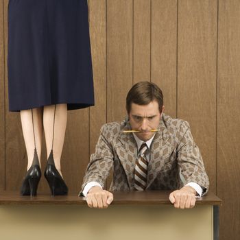 Mid-adult Caucasian male biting pencil and holding desk with Caucasian female standing on desk beside him.