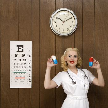 Caucasian mid-adult female nurse standing by wood paneling holding oversized pills and smiling at viewer.