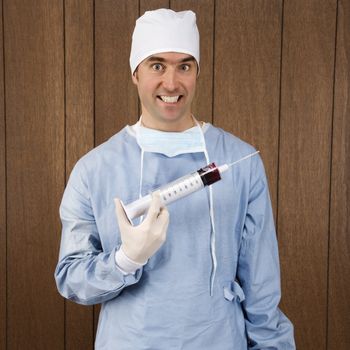 Mid-adult Caucasian male surgeon holding oversized syringe and smiling maniacally.