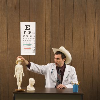 Mid-adult Caucasian male doctor wearing cowboy hat sitting at desk playing with figurine.