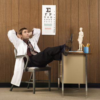 Mid-adult Caucasian male doctor at desk reclining with hands behind head and feet on desk.