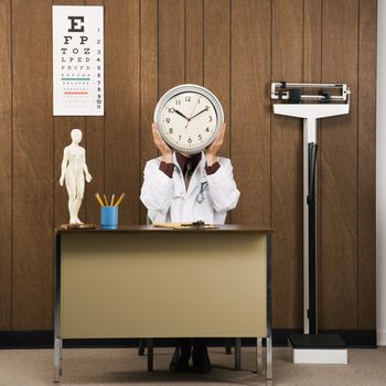 Caucasian male doctor sitting at desk holding clock over face.