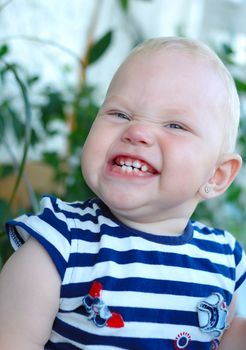 Little girl with blond hair smiling and grimacing on green plant background.