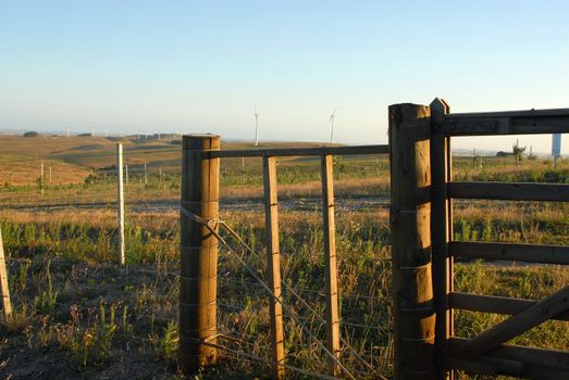 Wooden gate in wire fence at a windmills farm. Rural landscape under a blue sunny sky.