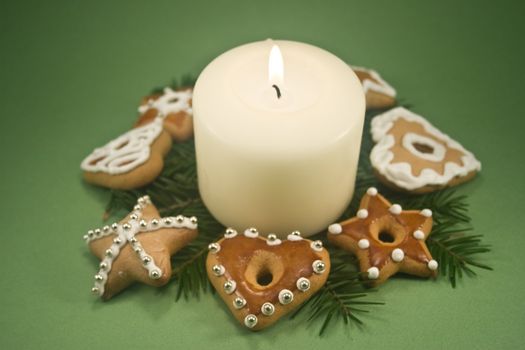 White pillar candle on fir branches surrounded by decorated gingerbread cookies on green background